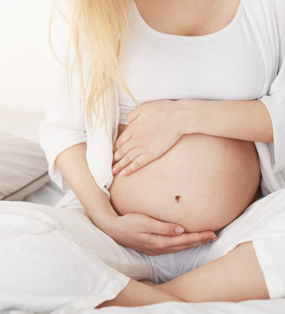 Young pregnant lady touching her belly sitting in her bedroom in afternoon. Pregnancy concept.